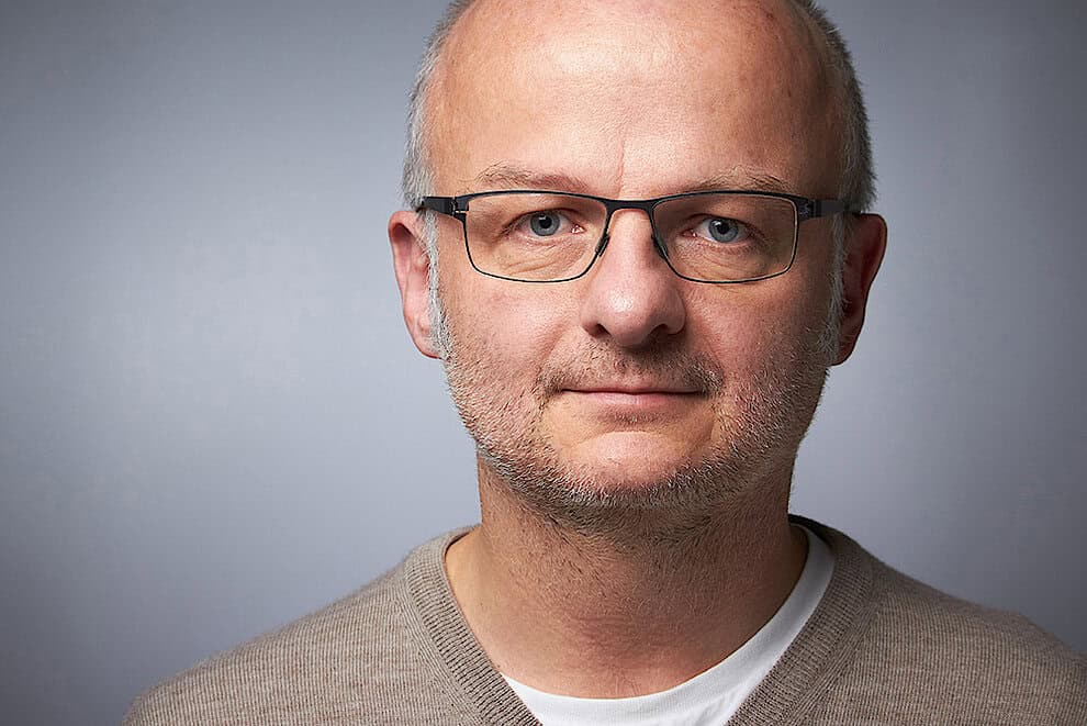 Ein Mann, der eine Brille und einen beigen Pullover traegt.